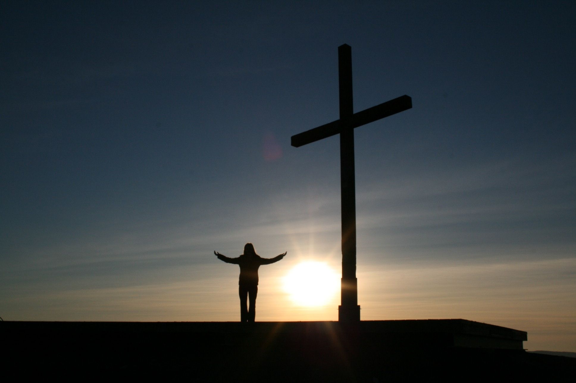 Silhouette of a person with arms raised standing beside a cross at sunrise